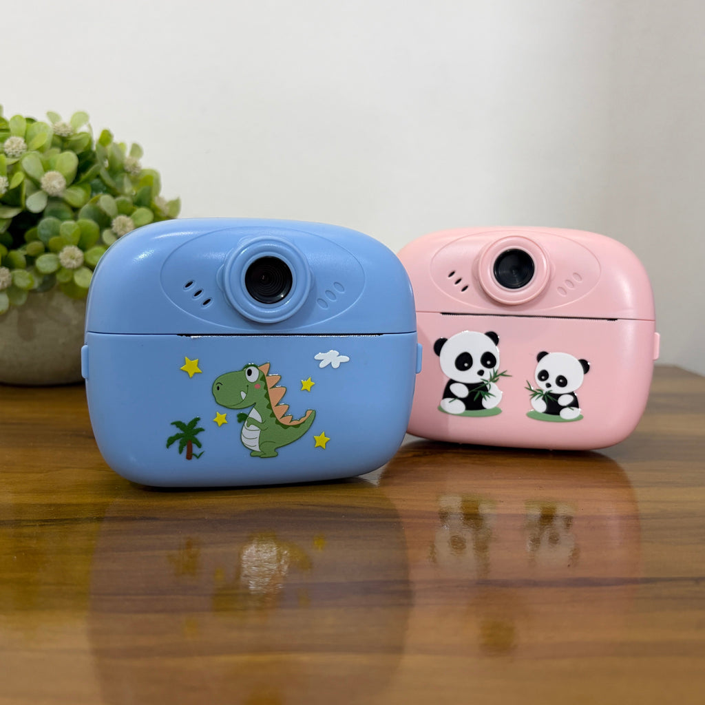 Two small compact cameras, one light blue with a green dinosaur and one light pink with two panda illustrations, displayed on a wooden table with a small potted plant in the background.