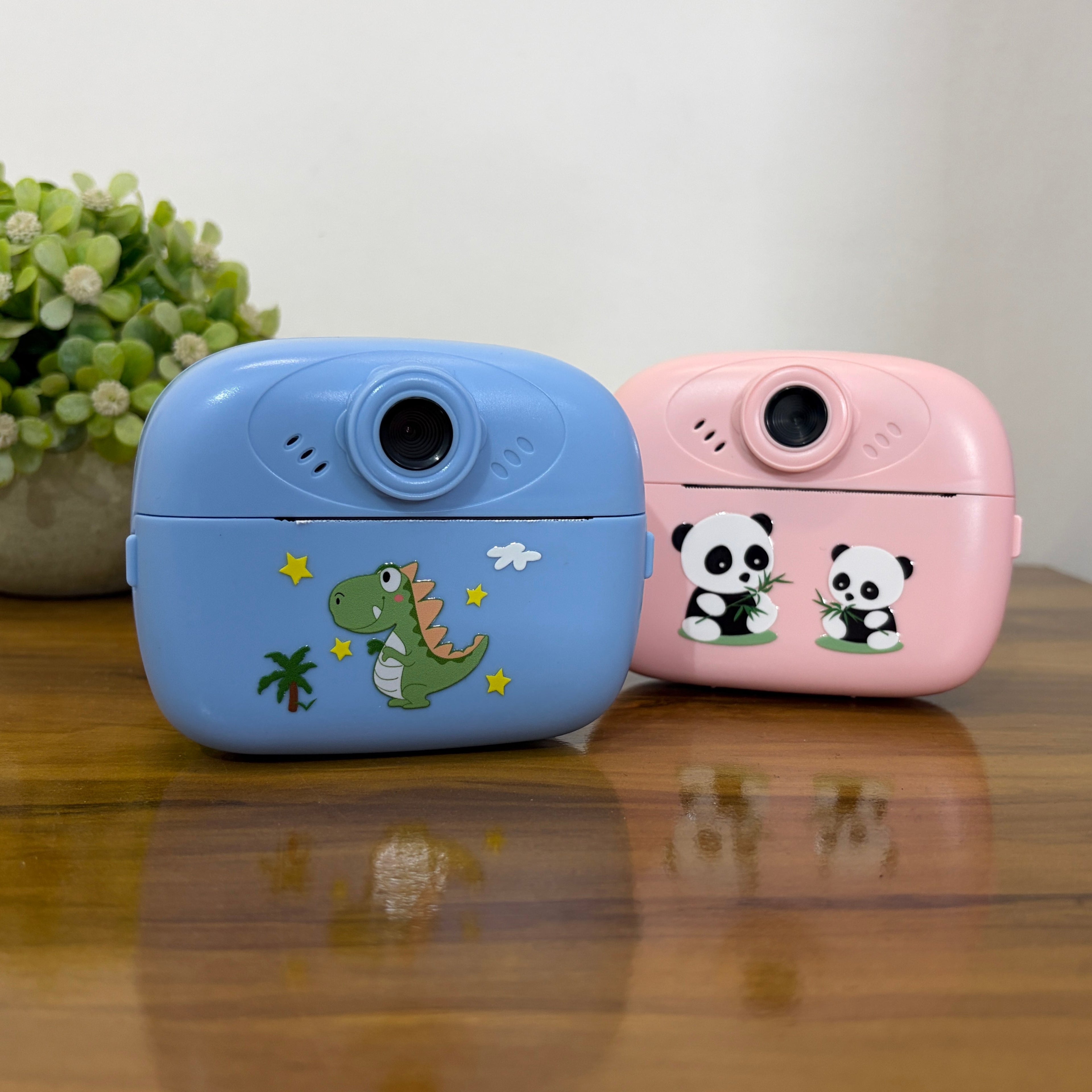 Two small compact cameras, one light blue with a green dinosaur and one light pink with two panda illustrations, displayed on a wooden table with a small potted plant in the background.