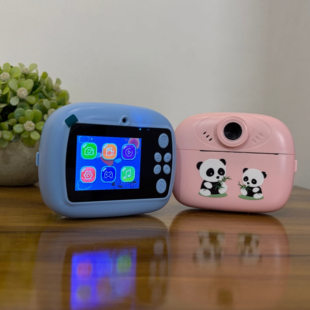 Two children's toy cameras, one light blue with a color screen and one pink with panda illustrations, displayed on a wooden table next to a small potted plant.